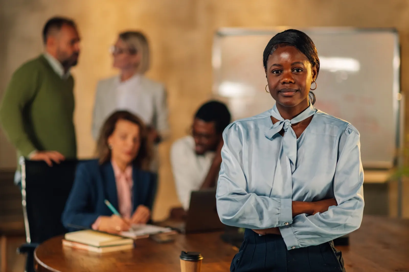 Professional standing with arms folded in front of colleagues meeting at a table